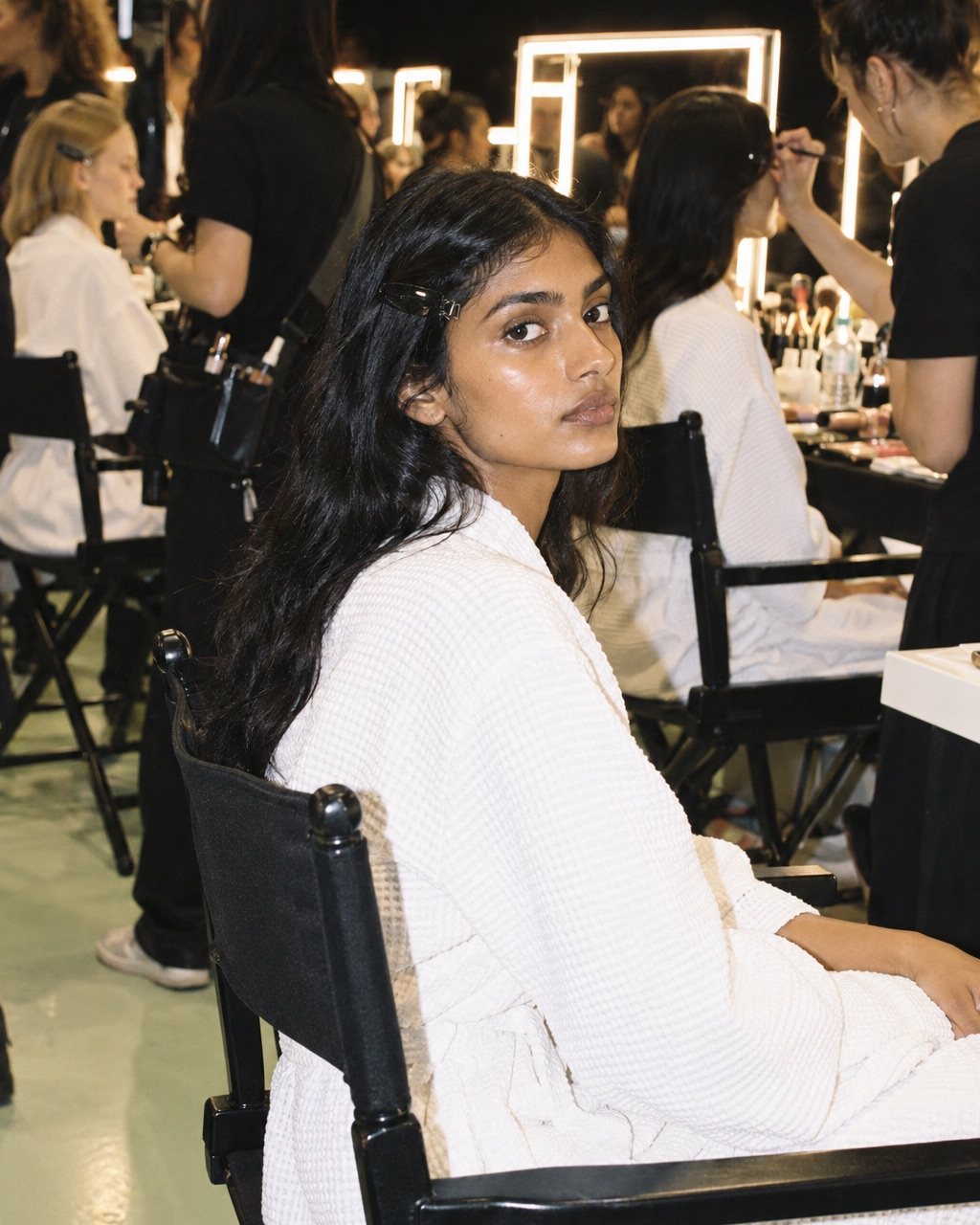 A model in a white robe sits backstage in a director's chair, skin prepped and bare